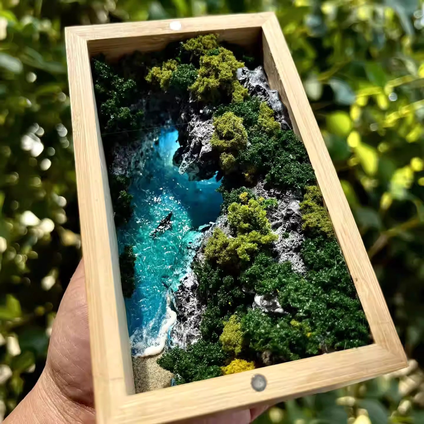 Framed artwork of a river surrounded by greenery held against a natural background