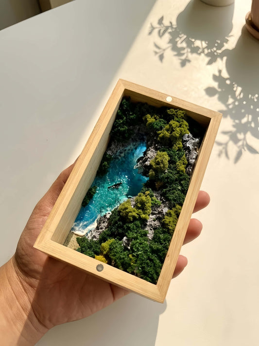 Hand holding a wooden-framed picture of a scenic landscape with trees and water.